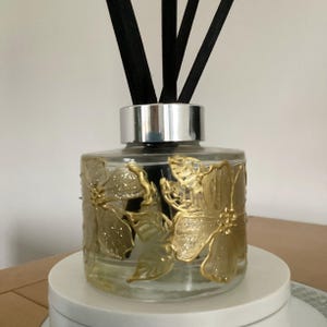 May include: A clear glass diffuser bottle with a silver lid and gold floral designs. Black diffuser sticks are in the bottle.