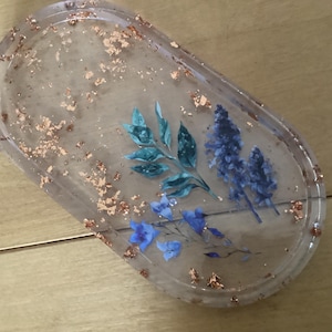 Puede incluir: Bandeja ovalada de resina con elementos florales azules y turquesas incrustados y copos dorados. La resina transparente permite ver las flores. Una pieza decorativa para joyas o objetos pequeños.