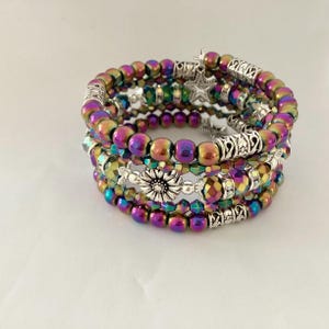 Könnte beinhalten: Ein mehrreihiges Wickelarmband mit irisierenden violetten, grünen und goldenen Perlen. Das Design umfasst silberfarbene Metallzwischenstücke, einen zentralen blumenförmigen Anhänger sowie einen kleinen sternförmigen Anhänger mit geschichteter Optik.