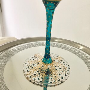 Op de afbeelding: Een handbeschilderd martiniglas met een turquoise steel en gouden en turquoise accenten. Het glas heeft een decoratief ontwerp op de kom en de voet, met een helder, reflecterend oppervlak. Het glas is ongeveer 20 cm hoog.