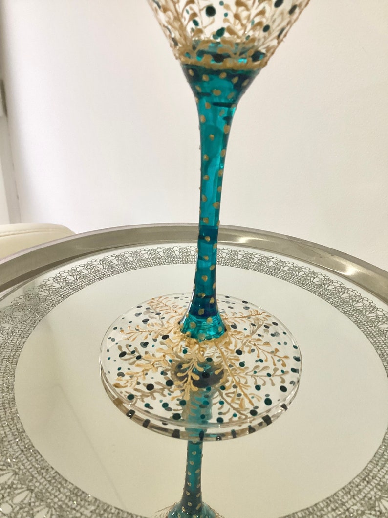Op de afbeelding: Een handbeschilderd martiniglas met een turquoise steel en gouden en turquoise accenten. Het glas heeft een decoratief ontwerp op de kom en de voet, met een helder, reflecterend oppervlak. Het glas is ongeveer 20 cm hoog.