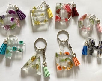 May include: Clear resin keychains in the shape of letters, each filled with dried flowers. The keychains have silver-colored hardware and colorful tassel accents. The letters include A, B, C, D, E, F, G, and H.