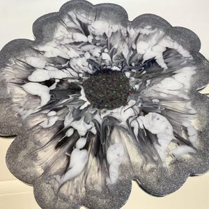 Puede incluir: Un posavasos de resina de color plateado y blanco con un diseño de mármol blanco y negro y un centro lleno de purpurina negra.