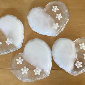 May include: Four heart-shaped coasters with a clear, glittery border and a white, marbled centre. Each coaster is adorned with small, white flower embellishments. The coasters are arranged on a light brown wooden surface.
