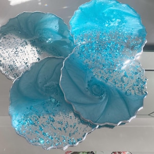 Puede incluir: Cuatro posavasos decorativos de resina azul claro con detalles de purpurina plateada y borde plateado. Presentan un diseño en espiral. Cada posavasos tiene un diámetro de aproximadamente 10 cm.