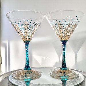 Op de afbeelding: Twee heldere martiniglazen met teal en gouden geschilderde ontwerpen. De stelen zijn teal met gouden stippen en de basis en de kom hebben gouden en teal blad- en stippatronen. De glazen staan op een zilveren dienblad.