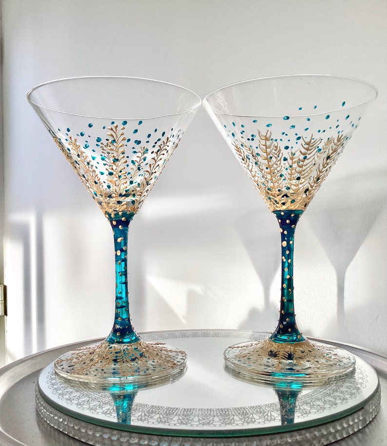 Op de afbeelding: Twee heldere martiniglazen met teal en gouden geschilderde ontwerpen. De stelen zijn teal met gouden stippen en de basis en de kom hebben gouden en teal blad- en stippatronen. De glazen staan op een zilveren dienblad.