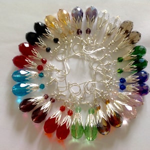 May include: A set of 18 teardrop-shaped crystal earrings with silver-toned settings. The earrings are arranged in a circle, with each earring featuring a different colour crystal.