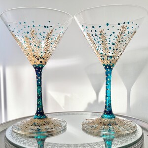 Op de afbeelding: Twee heldere martiniglazen met teal en goud geschilderde ontwerpen. De stelen zijn teal met gouden stippen en de bodems en kommen hebben ingewikkelde gouden en teal patronen. De glazen staan op een spiegelende tray.