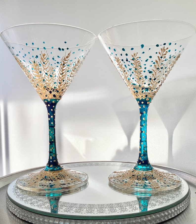 Op de afbeelding: Twee heldere martiniglazen met teal en goud geschilderde ontwerpen. De stelen zijn teal met gouden stippen en de bodems en kommen hebben ingewikkelde gouden en teal patronen. De glazen staan op een spiegelende tray.