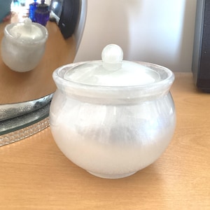 May include: A small, pearlescent white glass jar with a lid and a round knob on top. The jar is on a light brown wooden surface. A reflection of the jar is visible in a nearby mirror.