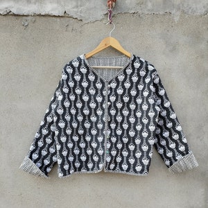 Puede incluir: Chaqueta estampada en blanco y negro con cuello en V y mangas largas. La chaqueta presenta un diseño floral blanco repetido sobre un fondo negro. Los puños y el forro interior tienen un estampado de rayas. La chaqueta está colgada en una percha de madera.