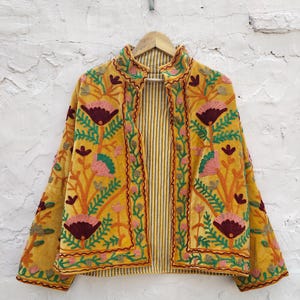 May include: Mustard yellow embroidered jacket with floral and leaf patterns in pink, green, and burgundy. The jacket has a striped lining and is hanging on a wooden hanger. The jacket's design is a blend of colours and textures.