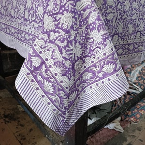 May include: A purple and white floral tablecloth draped over a table. The tablecloth has a repeating pattern of flowers and leaves. The border has a striped design. The fabric is folded over the table, showing the design.