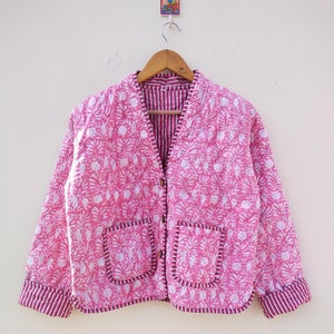 Puede incluir: Chaqueta acolchada rosa con un estampado floral blanco y dos bolsillos delanteros. La chaqueta tiene cuello en V y está forrada con una tela a rayas. Los puños y los bordes de los bolsillos están ribeteados con una tela a rayas.