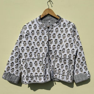 May include: White quilted jacket with a floral pattern in black and gold. The jacket has a mandarin collar, button closure, and black and white striped cuffs. The jacket is hanging on a wooden hanger.