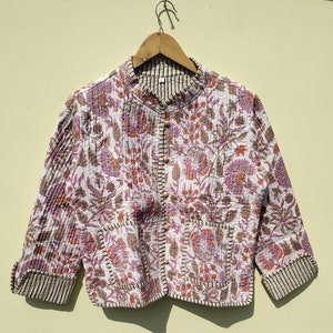 Puede incluir: Chaqueta acolchada con estampado floral en tonos blancos, rosas y marrones. Tiene cuello alzado, dos bolsillos y cierre de botones. Los puños y el cuello están ribeteados con una tela a rayas.