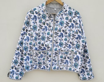 May include: A white quilted jacket with a blue floral pattern and black and white striped trim. The jacket has a round collar, two front pockets, and a full-length zipper. The jacket is hanging on a wooden hanger.