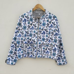 Puede incluir: Chaqueta acolchada blanca con estampado floral azul y ribete a rayas blancas y negras. La chaqueta tiene cuello redondo, dos bolsillos delanteros y cremallera completa. Cuelga de una percha de madera.