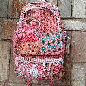 Puede incluir: Una mochila acolchada rosa con estampados florales en rojo, blanco y azul. La mochila tiene un asa superior, dos bolsillos delanteros con cremallera y correas ajustables. El diseño presenta una mezcla de patrones florales y geométricos.
