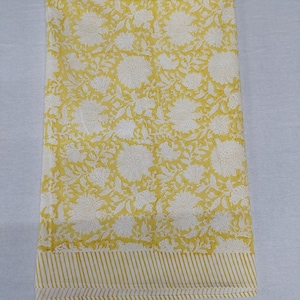 Op de afbeelding: Een gevouwen gele stof met een wit bloemenpatroon. De stof heeft een gestreepte rand aan de onderkant. Het bloemmotief heeft grote, gestileerde bloemen en bladeren. De algehele esthetiek is helder en vrolijk.