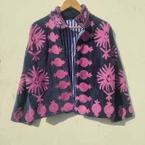 May include: A dark blue jacket with pink floral embroidery. The jacket has a striped blue and white lining and is hanging on a wooden hanger. The embroidery features stylized flowers and other shapes.