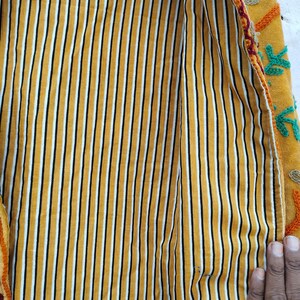 May include: A close-up shot of a yellow jacket's interior, featuring vertical black and white stripes. The jacket's exterior is mustard yellow with colourful floral embroidery. The jacket is open, displaying the striped lining.