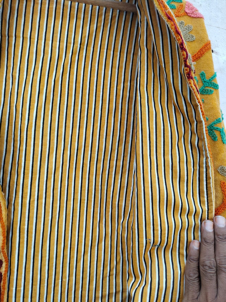 May include: A close-up shot of a yellow jacket's interior, featuring vertical black and white stripes. The jacket's exterior is mustard yellow with colourful floral embroidery. The jacket is open, displaying the striped lining.