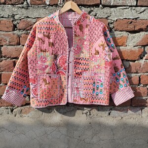 May include: A pink quilted jacket with a floral and geometric pattern. The jacket has long sleeves, a mandarin collar, and two front pockets. It is hanging on a wooden hanger against a brick wall.