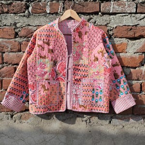 May include: A pink quilted jacket with a patchwork design. The jacket features floral and geometric patterns in shades of pink, red, and blue. It has a mandarin collar, long sleeves, and a button closure. The jacket is hanging on a wooden hanger against a brick wall.