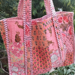Peut inclure: Sac fourre-tout matelassé avec un motif patchwork dans les tons rose et orange. Le sac présente des motifs floraux et tigrés, avec des poignées et des bords rayés. Fabriqué en tissu matelassé.