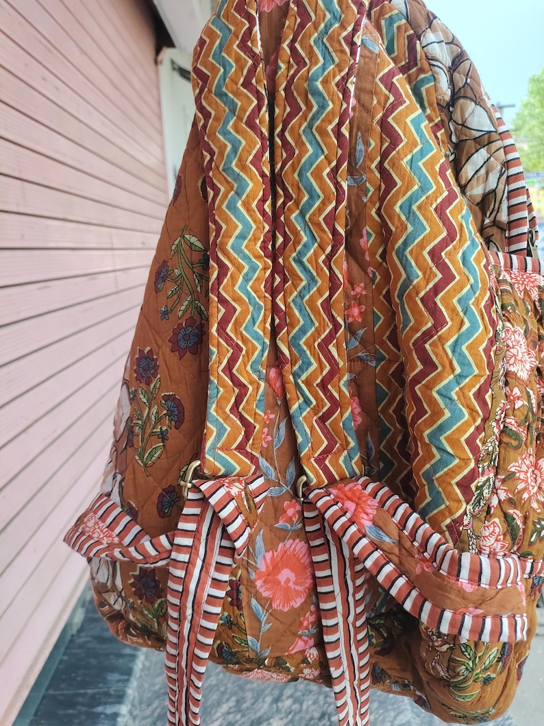 May include: Quilted backpack with a brown, orange, and teal zigzag pattern on the straps and a floral print on the body. The backpack has adjustable straps and a top handle. The bag is made of a soft, quilted fabric.