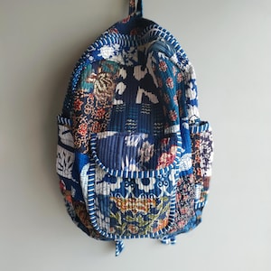 Op de afbeelding: Een gewatteerde rugzak met een patchworkontwerp van diverse blauwe, witte en veelkleurige bloemen- en geometrische patronen. De tas is voorzien van een voorvak met rits, zijvakken en een handvat aan de bovenkant, afgewerkt met verticale stiksels.