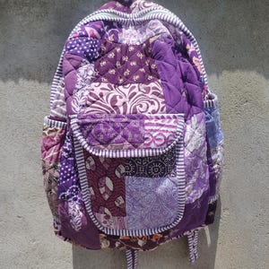 Pode incluir: Mochila acolchoada roxa, com um design de patchwork com vários padrões. A mochila tem um bolso frontal, bolsos laterais e uma alça superior. O design inclui padrões florais e geométricos.