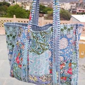 Puede incluir: Bolso tote acolchado con diseño patchwork. El bolso presenta varios estampados florales en tonos azules, turquesas y rosas. Las asas son a rayas azules y blancas. El bolso está hecho de tela acolchada.