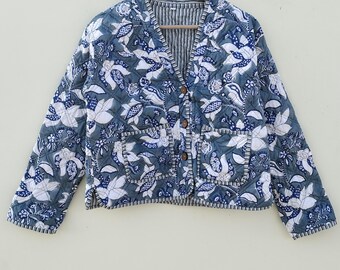 May include: A quilted jacket with a floral pattern in shades of blue and white. The jacket has a V-neck, wooden buttons, and two front pockets. It is hanging on a wooden hanger.