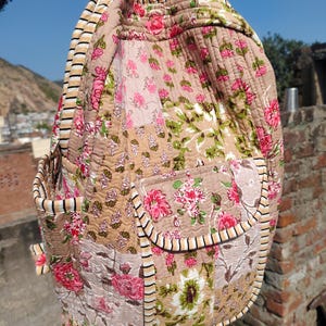 Op de afbeelding: Gewatteerde rugzak met een patchwork ontwerp met bloempatronen in roze, groen en beige. De rugzak heeft een afgeronde vorm, een voorvak en gestreepte rand. De tas is gemaakt van zachte, gewatteerde stof.