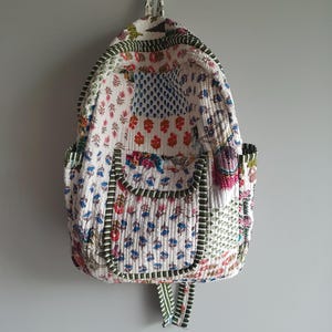 May include: A quilted backpack featuring a patchwork design with various floral and botanical patterns in shades of red, blue, green, and pink on a white background. The bag includes a front pocket, side pockets, and green striped trim.