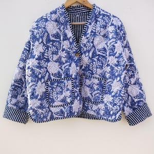 May include: A blue and white floral print jacket with two front pockets and a striped trim. The jacket is quilted and has a button closure. The jacket is hanging on a wooden hanger.