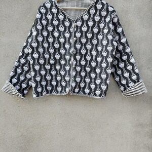 Puede incluir: Chaqueta estampada en blanco y negro con un diseño floral. La chaqueta tiene mangas largas y una botonadura frontal. Los puños y el cuello son a rayas. La chaqueta está colgada de una percha de madera.