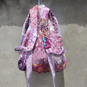 Könnte beinhalten: Ein gesteppter Rucksack mit einem Patchwork-Design in verschiedenen Violett-, Rosa- und Magentatönen. Die Tasche besteht aus mehreren Stoffmustern, darunter Blumen- und geometrische Drucke, mit gestreiften Stoffakzenten an den Schultergurten.