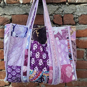 May include: A patchwork tote bag in shades of purple, lavender, and white, featuring floral and paisley patterns. The bag has matching purple handles with white trim. The bag is suspended against a brick wall. The bag's design is quilted.