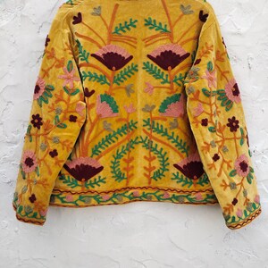 May include: Mustard yellow velvet jacket with detailed floral embroidery in pink, burgundy, green, and gold. The jacket has a high collar and long sleeves, displaying intricate craftsmanship. The floral design is symmetrical, covering the back and sleeves.