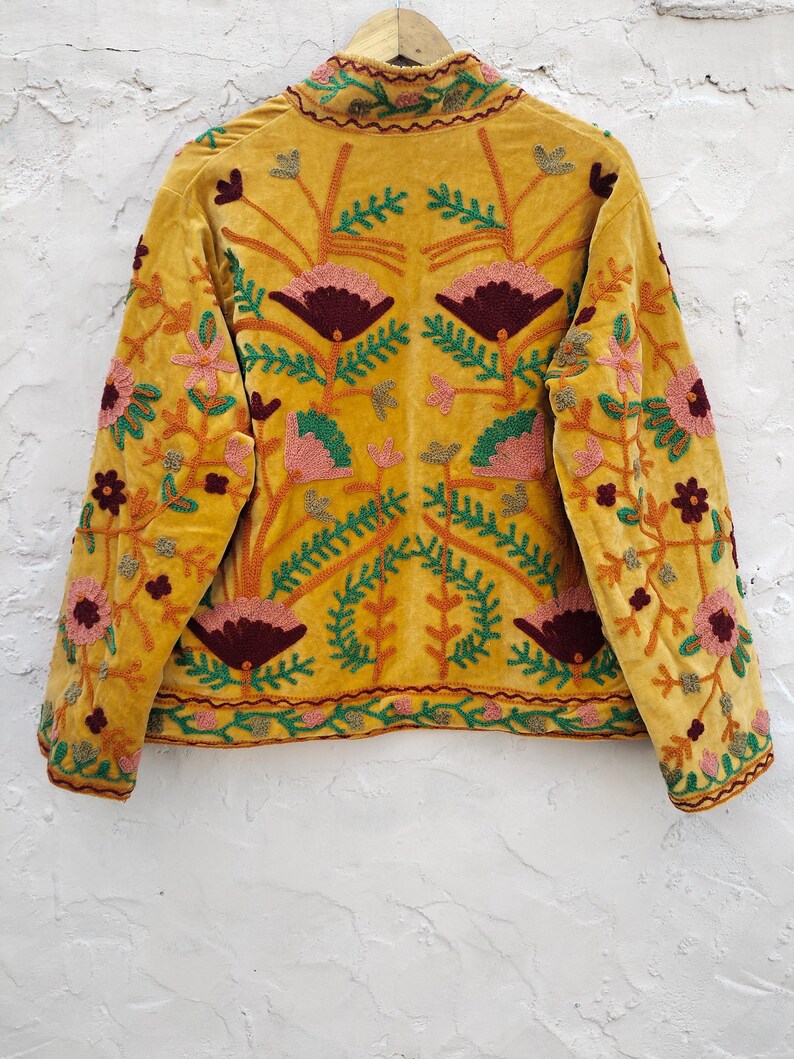 May include: Mustard yellow velvet jacket with detailed floral embroidery in pink, burgundy, green, and gold. The jacket has a high collar and long sleeves, displaying intricate craftsmanship. The floral design is symmetrical, covering the back and sleeves.