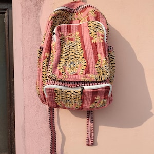 Könnte beinhalten: Ein gesteppter Rucksack mit einem sich wiederholenden Tigermuster in Rosa-, Gelb- und Schwarztönen. Die Tasche verfügt über ein großes Hauptfach, eine kleinere Vordertasche und zwei Seitentaschen, die alle mit weißen Reißverschlüssen versehen sind.