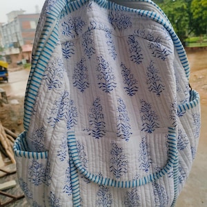 May include: White quilted backpack with a blue floral design. The backpack has blue striped edging and two side pockets. The bag is made of a soft, quilted fabric and features a top zip closure.
