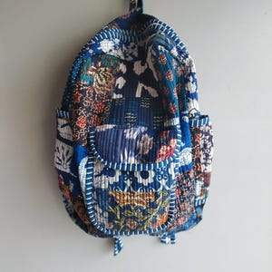 Puede incluir: Una mochila acolchada con un diseño de patchwork que combina varios patrones de tela florales y geométricos en tonos azul, blanco y multicolor. La bolsa incluye un bolsillo frontal con cremallera, dos bolsillos laterales y un asa superior.