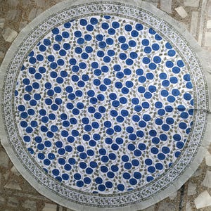 Könnte beinhalten: Eine runde Tischdecke mit weißem Hintergrund und einem sich wiederholenden Muster aus blauen, kreisförmigen Blumenmotiven und grünen Ranken. Der Rand besteht aus einem dekorativen Blumenband und einem schmalen, hellen Streifen.