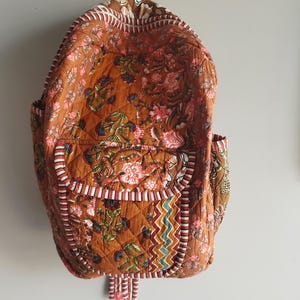 May include: Quilted backpack with a floral pattern in shades of brown, pink, and green. The backpack has two side pockets and a front pocket with a decorative trim. The straps and handle are brown.