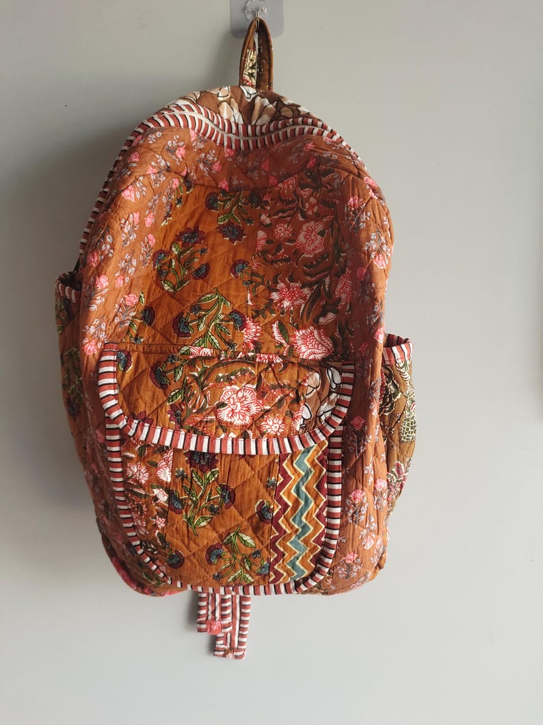 May include: Quilted backpack with a floral pattern in shades of brown, pink, and green. The backpack has two side pockets and a front pocket with a decorative trim. The straps and handle are brown.